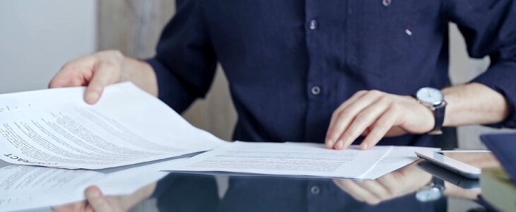 Businessman in dark blue t-shirt analyzing document at desk. Close-up of a professional auditor or lawyer reviewing a lengthy paper report in office setting. Business people concept.